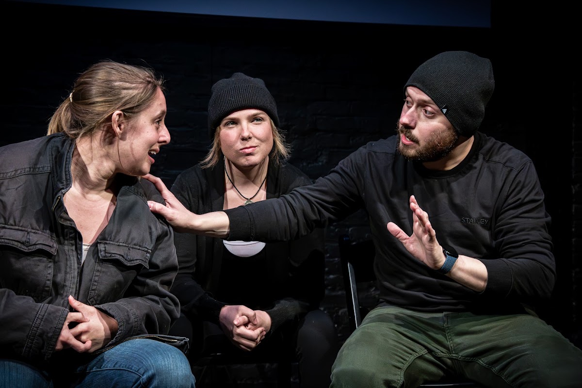 Three performers crowded knee to knee, hands up mid-argument.